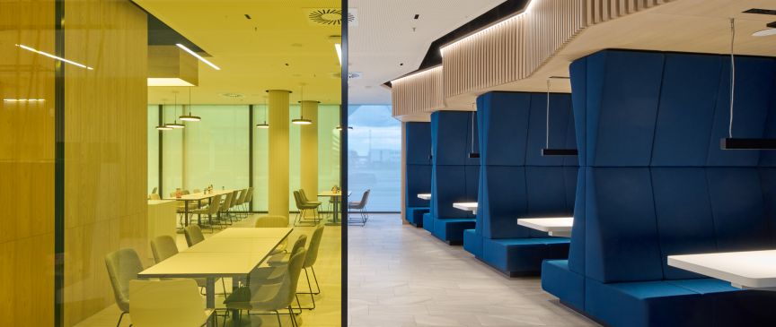 Modernes Büro: links gelb verglaster Meetingraum mit Tisch und Stühlen, rechts blaue Sitzkojen mit weißen Tischen.