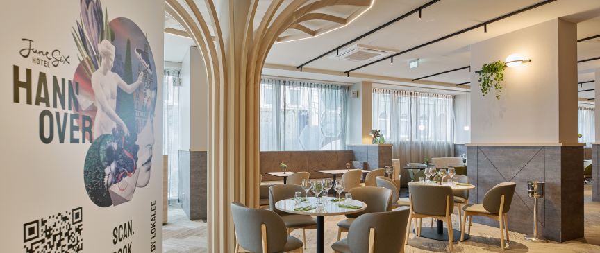 Modernes Hotelrestaurant mit runden Tischen, grauen Stühlen, dekorativen Holzsäulen und einem Banner mit "Hannover" und QR-Code.