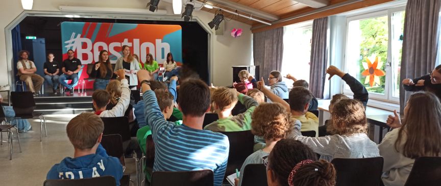 Kinder sitzen in einem Saal und strecken die Hand auf. Auf einer Bühne mit "#Bestjob"-Banner sitzen mehrere Erwachsene.