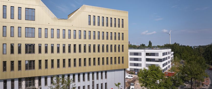 Modernes Bürogebäude mit goldfarbener Fassade im Bau, dahinter weißes Gebäude, Windrad und Bäume unter blauem Himmel.