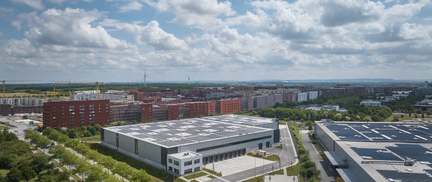 Luftaufnahme eines Industriegebiets mit Lagerhallen und Solardächern, angrenzend an Wohngebäude und Grünflächen.