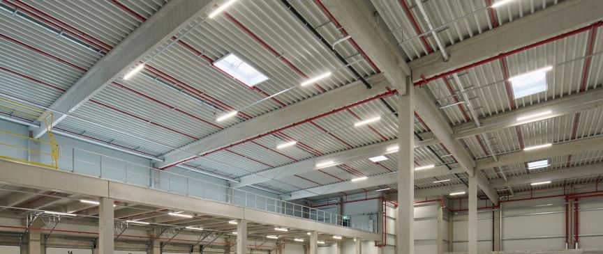 Leeres Industrielager mit Betonboden, Stahlträgern, roten Rohren an der Decke und mehreren Garagentoren an der Wand.