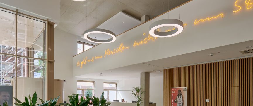Modernes Foyer mit runden Deckenleuchten, Neonschrift auf Deutsch, Holzwandverkleidung, Pflanzen und großen Fenstern.
