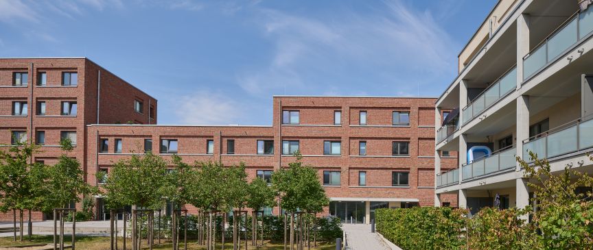 Innenhof mit Picknickbänken, Bäumen und Gehweg zwischen modernen Backsteingebäuden mit Balkonen unter blauem Himmel.