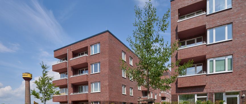 Zwei mehrstöckige Backsteingebäude mit Balkonen, junge Bäume im Innenhof, blauer Himmel, gelber Wasserturm im Hintergrund.