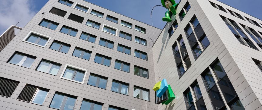 Modernes Bürogebäude mit grauer Fassade, vielen Fenstern, grüner Ampel und buntem Logo im Vordergrund, blauer Himmel.