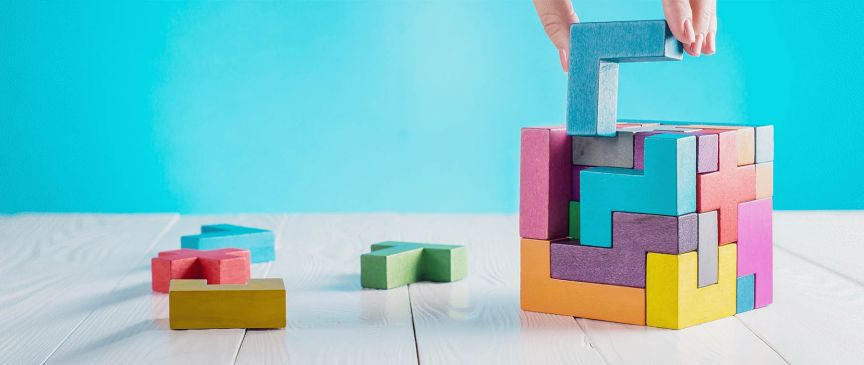 Hand platziert blauen L-förmigen Holzblock auf bunten Tetris-ähnlichen Holzblöcken auf weißem Tisch.