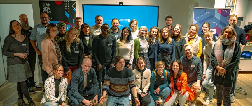 Gruppe von ca. 25 Personen posiert für ein Gruppenfoto in einem modernen Büroraum mit großem Bildschirm.