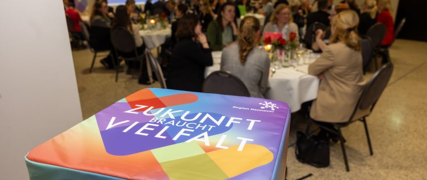 Bunte Sitzwürfel mit Aufschrift „Zukunft braucht Vielfalt" im Vordergrund, dahinter Veranstaltungsraum mit Publikum an runden Tischen.