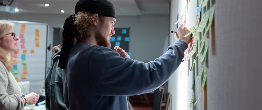 Mann mit Bart und Mütze schreibt mit Marker auf Post-its an einer Wand. Zwei Personen stehen daneben.