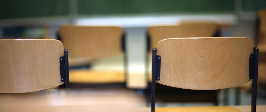 Leere Holzstühle in einem Klassenzimmer mit grüner Tafel im Hintergrund.