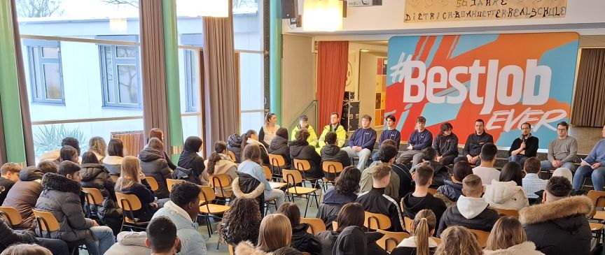 Schüler sitzen in Stuhlreihen vor einem Panel mit Erwachsenen in einer Aula. Banner mit "#BestJob Ever" im Hintergrund.