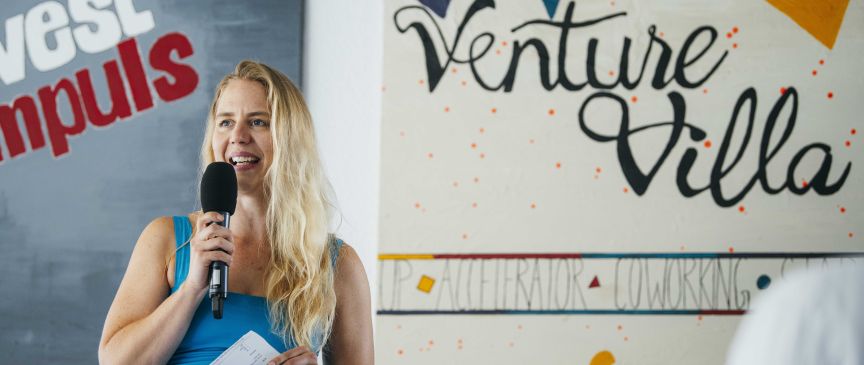 Blonde Frau mit Mikrofon spricht vor einem Venture Villa Schild, hält Papier in der Hand.