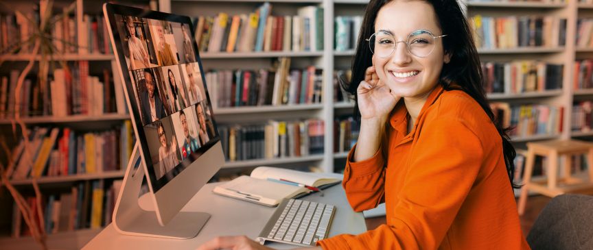 Junge Frau mit Brille und orangem Pullover sitzt vor einem iMac mit Videokonferenz in einer Bibliothek.