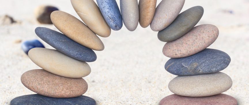 Zwei Steinstapel auf Sand, verbunden durch einen Bogen aus flachen, ovalen Kieseln in verschiedenen Farben.