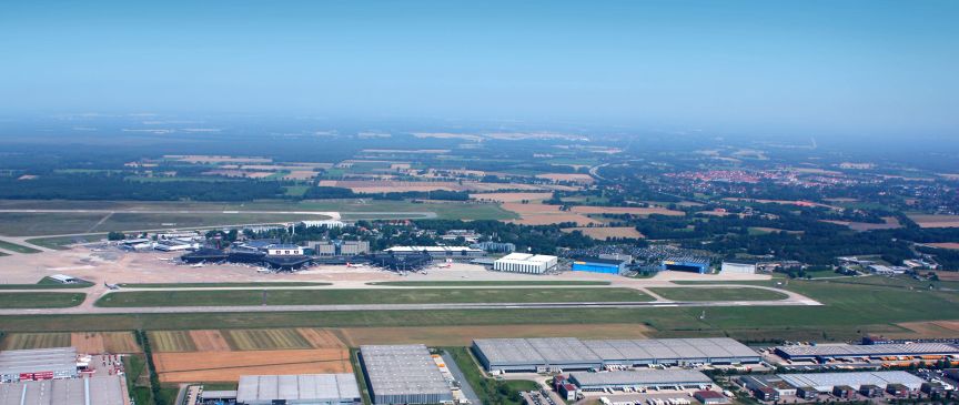Luftaufnahme eines Flughafens mit Landebahn, Terminals und Hangars, umgeben von Feldern und Industriegebäuden.