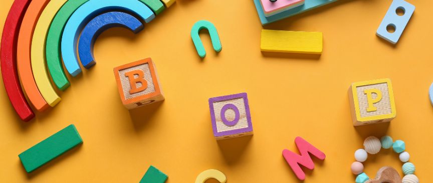 Bunte Holzspielzeuge auf orangem Hintergrund: Regenbogen, Buchstabenblöcke (B, O, P, M, C, S, J), Bausteine und Beißring.