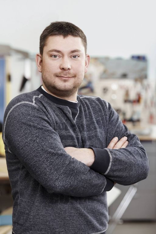 Mann mit kurzen Haaren, verschränkten Armen, grauem Pullover, steht in einer Werkstatt.