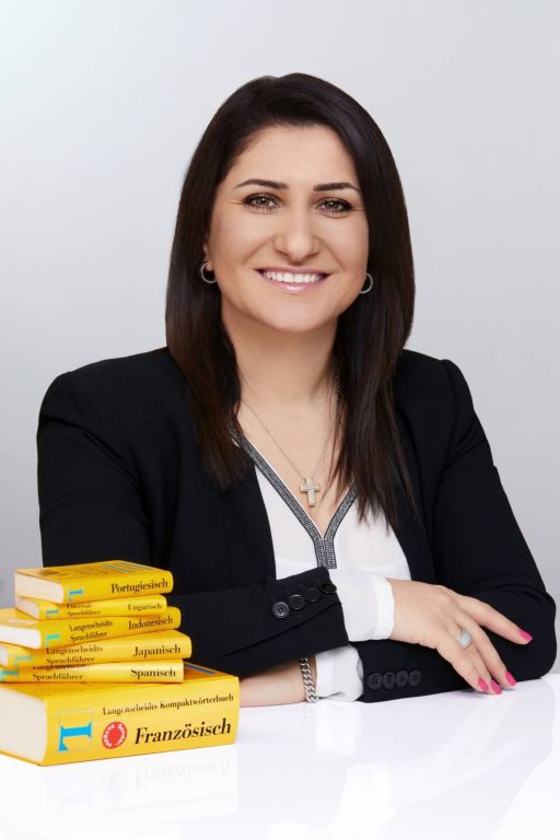 Frau in schwarzem Blazer sitzt an einem Tisch mit einem Stapel Langenscheidt-Wörterbücher auf Deutsch.