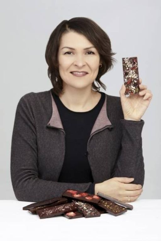 Frau hält Schokoladenriegel hoch, vor ihr liegen mehrere Schokoladentafeln auf weißem Tisch.