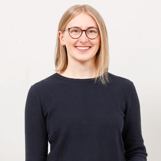Lächelnde Frau mit blonden Haaren und Brille in dunklem Pullover vor hellem Hintergrund.