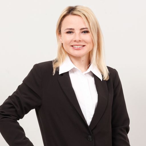 Blonde Frau im schwarzen Blazer mit weißem Hemd lächelt, Hand in der Hüfte, heller Hintergrund.
