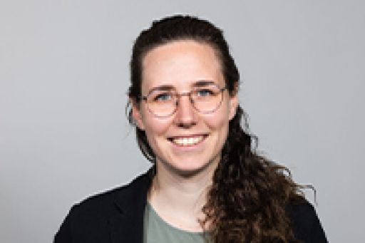 Lächelnde Person mit Brille und lockigem braunem Haar, schwarzer Jacke, vor grauem Hintergrund.