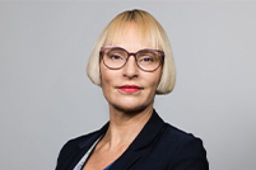 Frau mit blondem Bob, Brille und dunklem Blazer vor grauem Hintergrund, Porträtfoto.