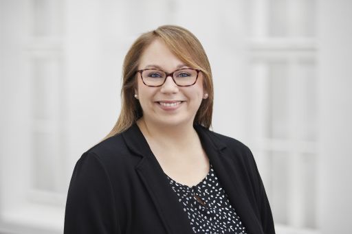 Frau mit brauner Brille und langen rötlich-braunen Haaren im schwarzen Blazer lächelt vor hellem Hintergrund.