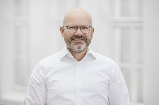Mann mit Glatze, Brille und grauem Bart, weißes Hemd, lächelt vor hellem Hintergrund.