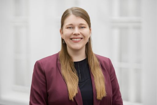 Junge Frau mit langen blonden Haaren, rotem Blazer und schwarzem Oberteil lächelt vor hellem Hintergrund.