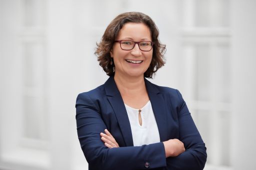 Frau mit Brille und dunklem Blazer, Arme verschränkt, lächelt vor hellem Hintergrund.