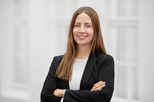 Frau mit langen braunen Haaren, schwarzem Blazer, verschränkten Armen, lächelnd vor hellem Hintergrund.