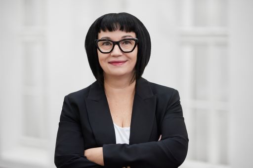 Frau mit schwarzem Bob, Brille und Blazer, Arme verschränkt, lächelt vor hellem Hintergrund.
