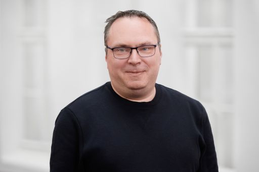 Mann mit Brille und kurzem Haar trägt schwarzen Pullover, lächelt leicht, vor hellem Hintergrund.