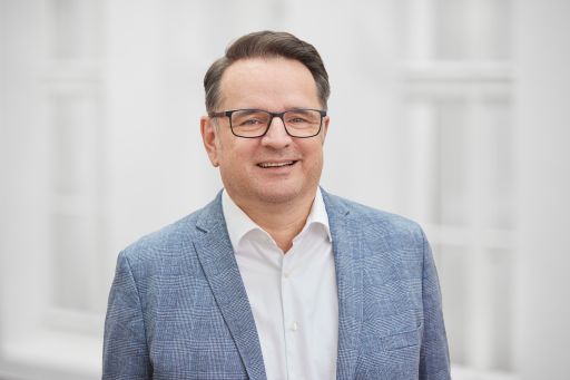Mann mit Brille und grauem Sakko lächelt vor hellem Hintergrund. Weißes Hemd, dunkles Haar.
