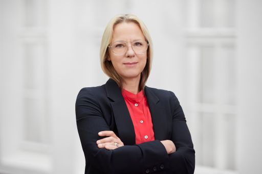 Frau mit blonden Haaren und Brille, schwarzem Blazer und roter Bluse, Arme verschränkt, vor hellem Hintergrund.