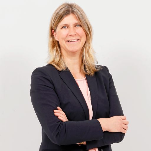 Frau mit blondem Haar, dunklem Blazer und rosa Bluse, Arme verschränkt, lächelt vor hellem Hintergrund.