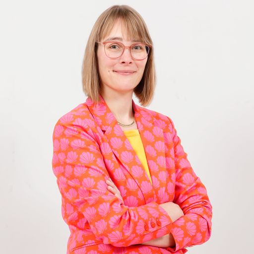 Frau mit Brille und schulterlangem Haar, in orangefarbenem Blazer mit rosa Muster, Arme verschränkt, vor weißem Hintergrund.