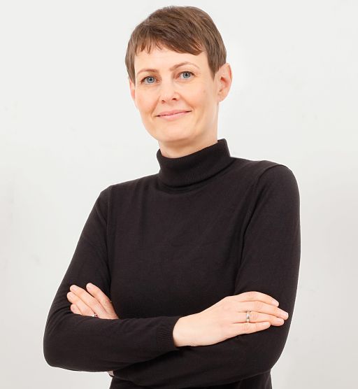 Frau mit kurzen braunen Haaren, schwarzem Rollkragenpullover, verschränkten Armen, lächelt vor hellem Hintergrund.