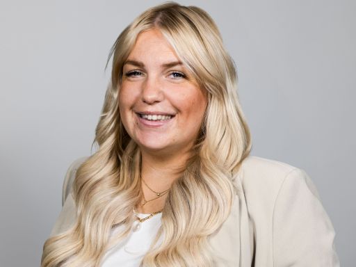 Lächelnde Frau mit langen blonden Haaren, beigem Blazer und Goldkette vor grauem Hintergrund.
