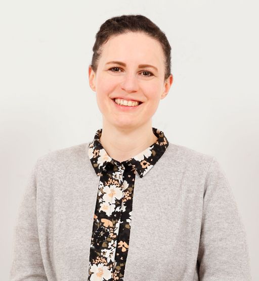 Lächelnde Frau mit dunklem Haar, grauem Cardigan und schwarz-weißer Bluse mit Blumenmuster vor hellem Hintergrund.
