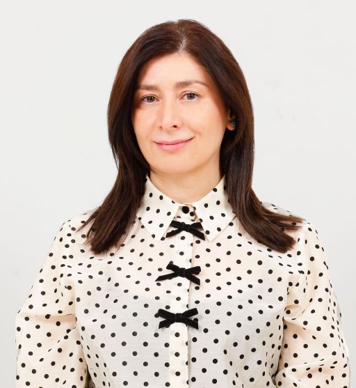 Frau mit schulterlangem braunem Haar in cremefarbenem Polka-Dot-Hemd mit schwarzen Schleifen, vor grauem Hintergrund.
