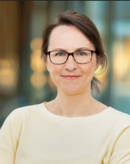 Frau mit dunklem Haar und Brille in cremefarbenem Oberteil lächelt in die Kamera, unscharfer Hintergrund.
