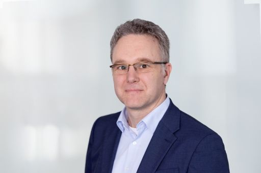 Mann mit Brille und grau-braunem Haar, blauem Sakko und hellblauem Hemd, vor hellem Hintergrund.