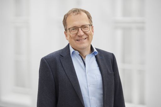 Mann mit Brille, grauem Haar, blauem Sakko und hellblauem Hemd lächelt vor weißem Hintergrund.