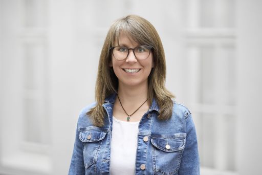 Frau mit Brille und schulterlagen braunen Haaren, Jeanjacke und Anhänger-Halskette lächelt in die Kamera.