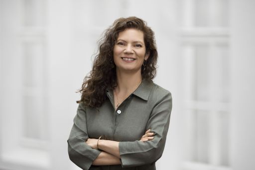 Frau mit lockigem braunem Haar, verschränkten Armen, grauem Hemd, lächelnd vor hellem Hintergrund.