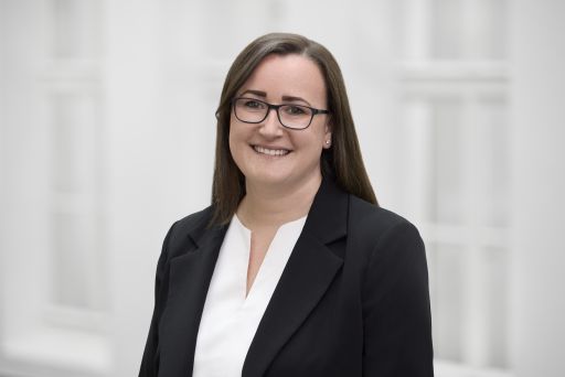 Frau mit braunen Haaren und Brille, schwarzem Blazer und weißer Bluse, lächelt in die Kamera.