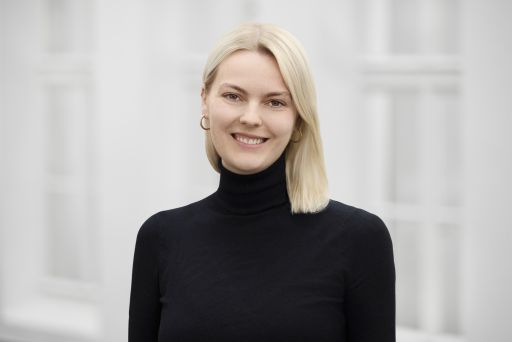 Junge Frau mit blonden Haaren und schwarzem Rollkragenpullover lächelt vor hellem Hintergrund.